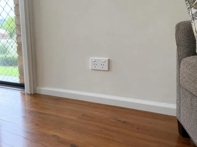 Double power point on a painted interior wall beside timber flooring and a living room chair in Ormeau