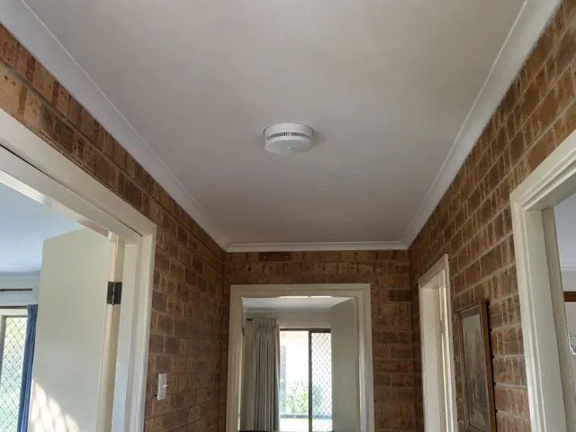Ceiling-mounted smoke alarm in a brick hallway with white trim in Coomera